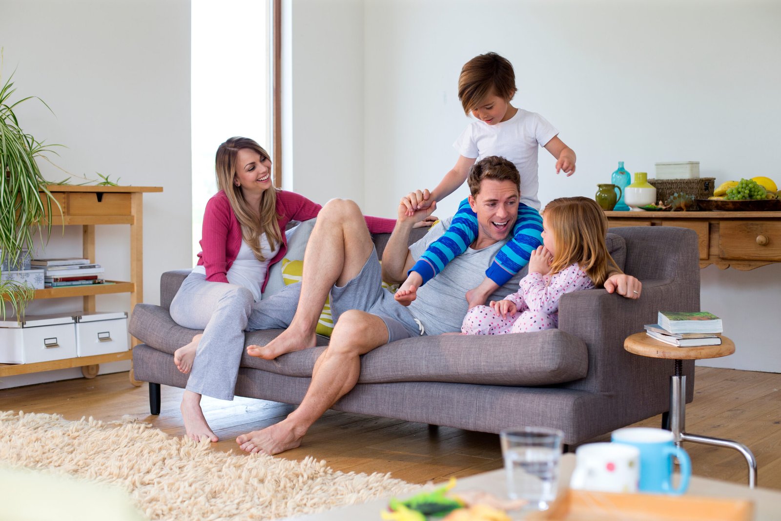 Family of four are sitting on the sofa in the living room of their home together. The father has his son on his shoulders and the little girl is sitting next to him.